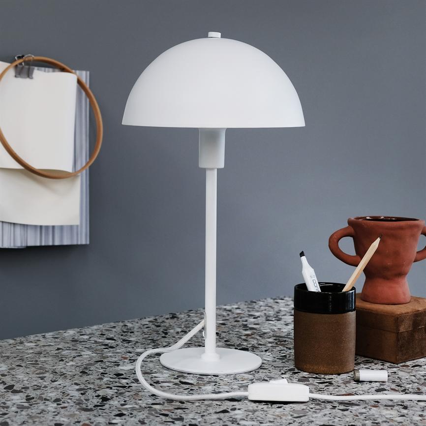 En hvid bordlampe står på et terrazzo-bord; dens kuppel sidder på en slank stok. Baggrunden er en grå væg med papirer og en broderiramme; til højre penneholder og terrakottakrukke.