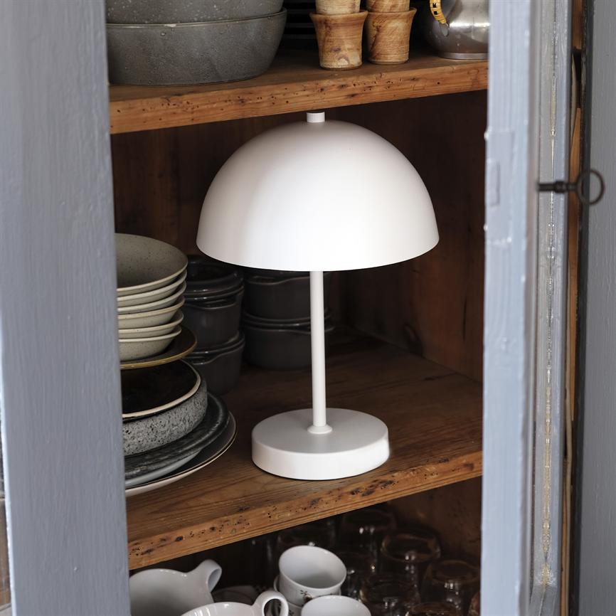 Hvid, halvkugleformet bordlampe står på en træhylde omgivet af stabler af tallerkener, skåle, krus og glas inde i et åbent køkkenskab.