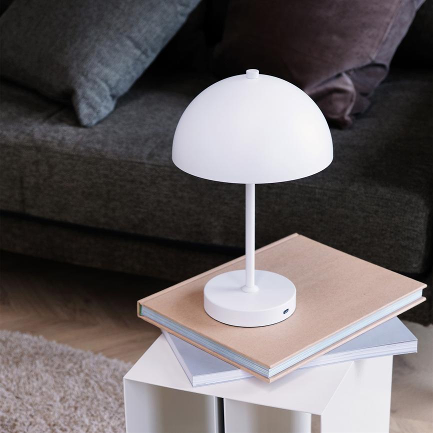 Hvid, halvrund bordlampe står slukket på en stak mapper på et lille hvidt sidebord; mørkegrå sofa og puder i baggrunden, lyst tæppe og trægulv under.
