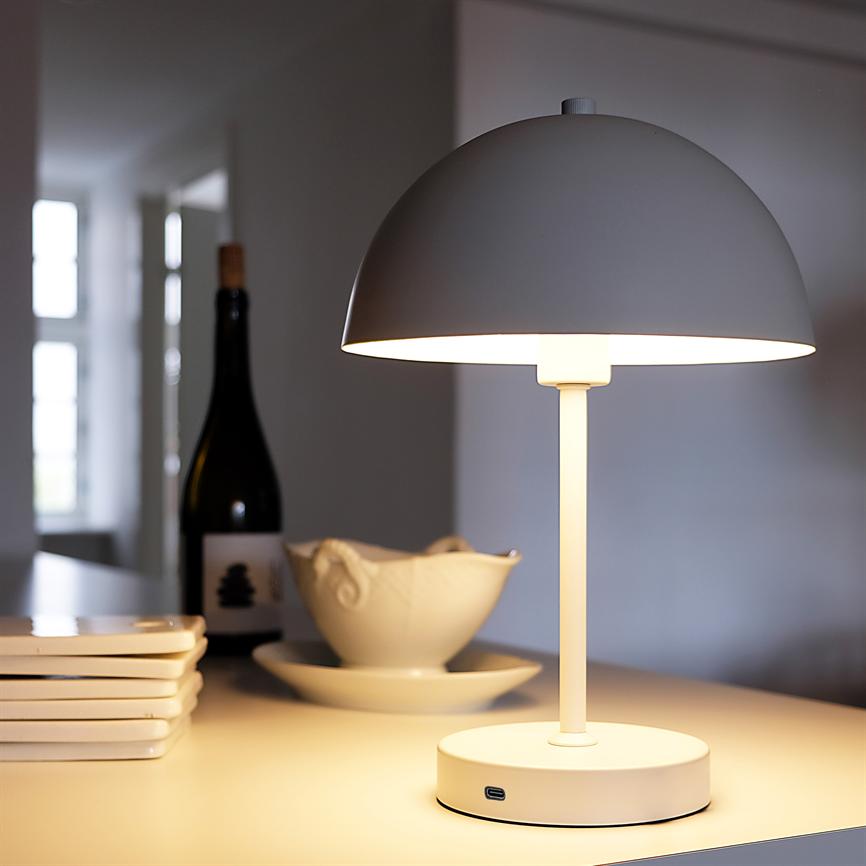 Bordlampe lyser på bordet; omgivet af en stak tallerkener, en saucekande og en vinflaske i et lyst, roligt køkkenmiljø.