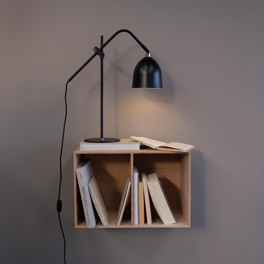 En sort arkitektlampe lyser over en åben bog på en lille træhylde monteret på en grå væg; hyldens rum rummer flere bøger. Tekst: "NOSTALGIA" (på en bogryg).