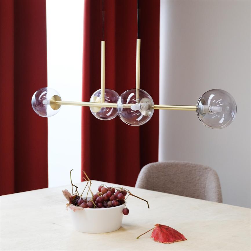 En moderne guldfarvet loftlampe med fire klare glaskupler hænger over et lyst spisebord; en hvid skål med røde druer står på bordet foran dybrøde gardiner og en blød stol.