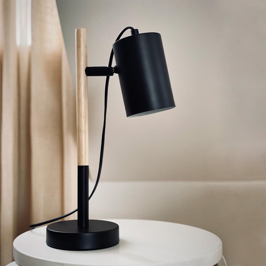 En sort og træ bordlampe står på et rundt hvidt bord; lampehovedet er vinklet nedad, ledningen hænger; baggrund: beige væg og lyse gardiner.