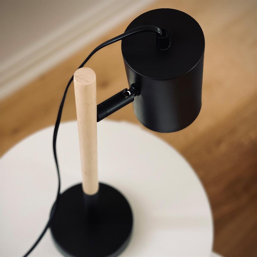 Bordlampe med sort metalskærm og lys træstang; kablet løber ned ad stangen; står på et hvidt rundt bord i et rum med varmt trægulv.