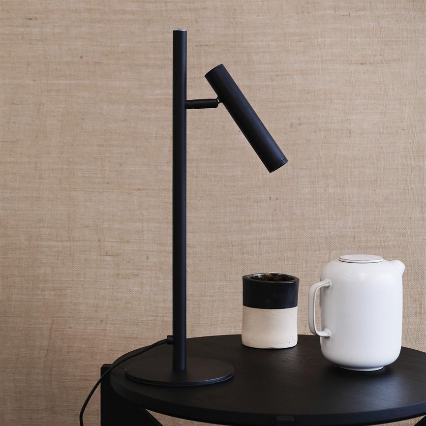 Sort bordlampe med cylindrisk hoved peger skråt nedad; står på rundt sort bord ved siden af en hvid tekande og en tofarvet kop; beige stofbaggrund.
