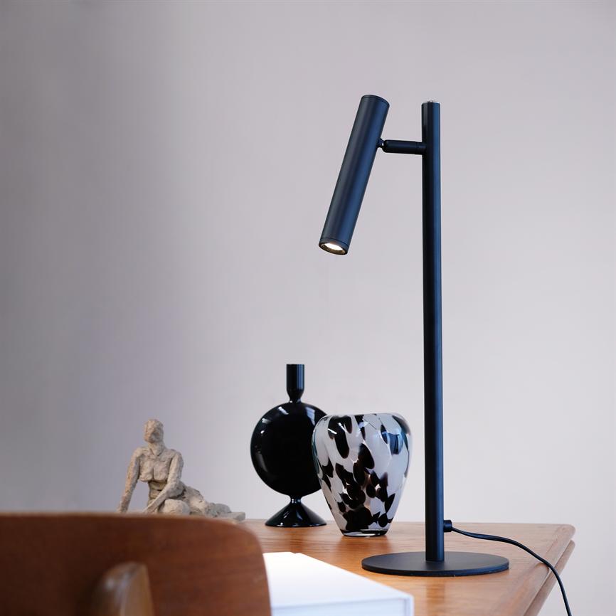 Sort, slank bordlampe kaster et smalt, varmt lys nedad på et træbord ved siden af en sort vase, en sort-hvid plettet vase og en lille skulptur foran en lys væg.