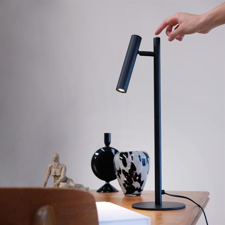 En sort, slank skrivebordslampe bliver rørt/tændt af en fingerspids, står på et træbord ved siden af to dekorative vaser og en lille skulptur i et lyst, neutralt rum.