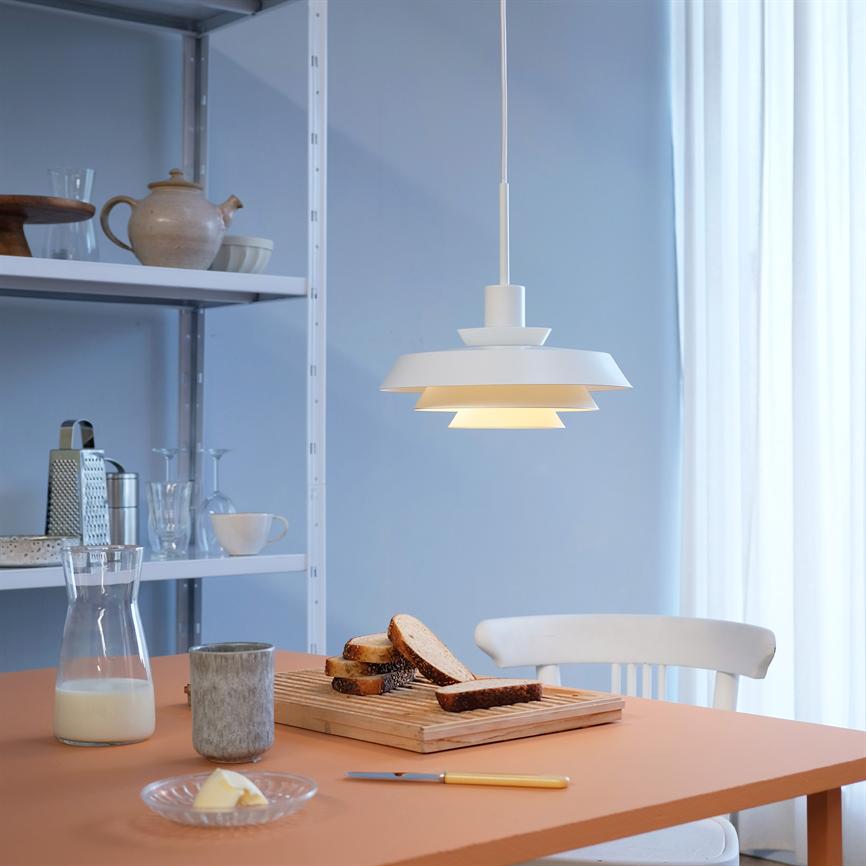 En hvid pendel lyser varmt over et køkkenbord, hvor et skærebræt med skiver af brød står med mælkekande, smør, krus, hylde med service og en hvid stol ved gardin.