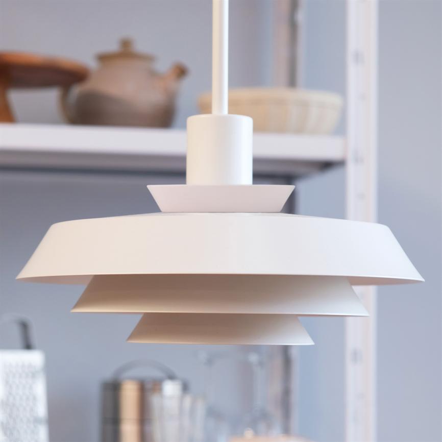 Hvid, lagdelt loftlampe hænger over et køkkenområde; baggrunden viser sløret hylde med keramikkrukker, skåle og glas.