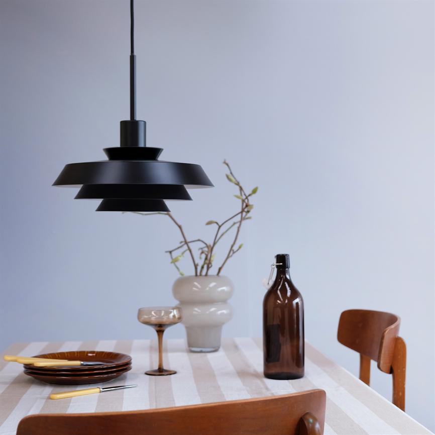 Sort, lagdelt loftslampe hænger over et stribet, dugebelagt spisestuebord med to brune stole; bordet rummer brune tallerkener, bestik, et glas, en brun flaske og en vase med grene.