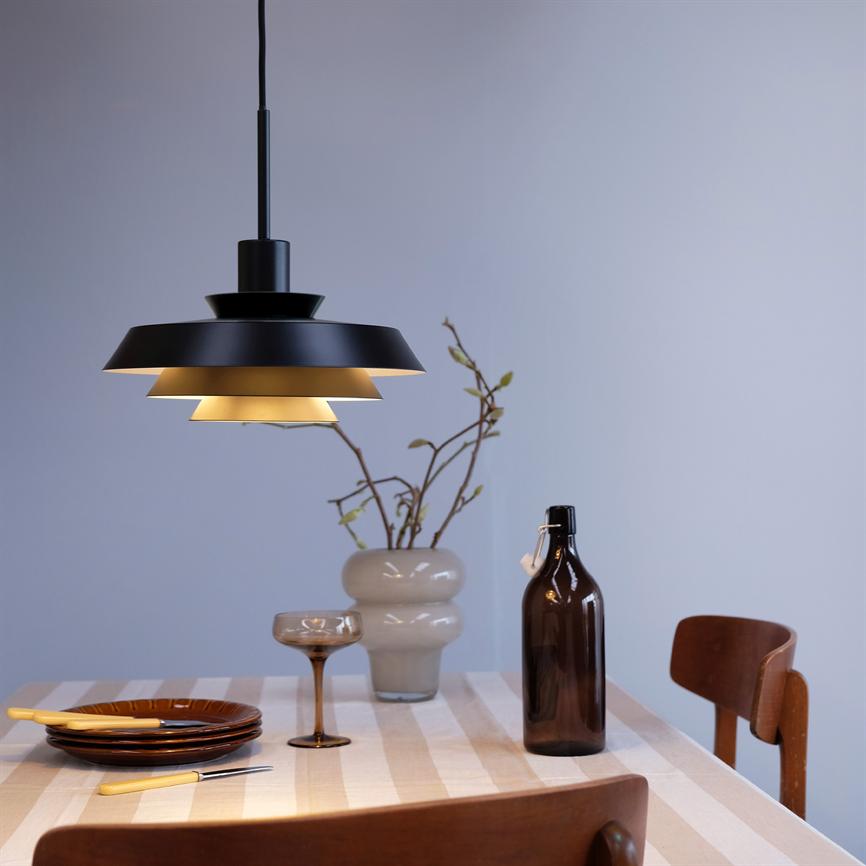Sort lagdelt loftslampe kaster varmt lys over et stribet dækket bord med tallerkener, cocktailglas, brun flaske og vase med grene i et lyst, enkelt spiserum.