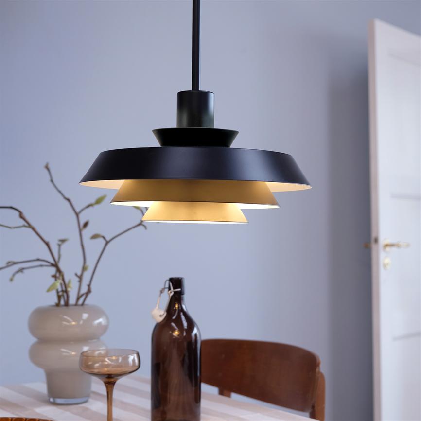 En sort, lagdelt pendel hænger og kaster varmt lys over et spisebord; på bordet står en vase med kviste, en brun flaske og et glas, bagved ses en åben dør.