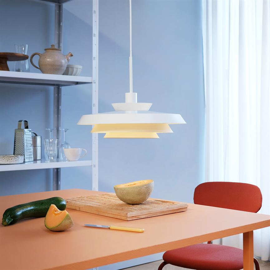 En hvid pendel lyser over et orange bord; en melonhalvdel på et skærebræt, en zucchini ved siden af, med rød stol og hylde i baggrunden.