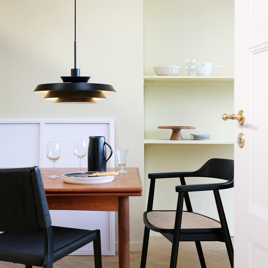 En sort pendellampe hænger og kaster varmt lys ned over et træspisebord; glas, en sort kande og tallerken ligger på bordet, omgivet af sorte stole og lysegule hylder med keramik.