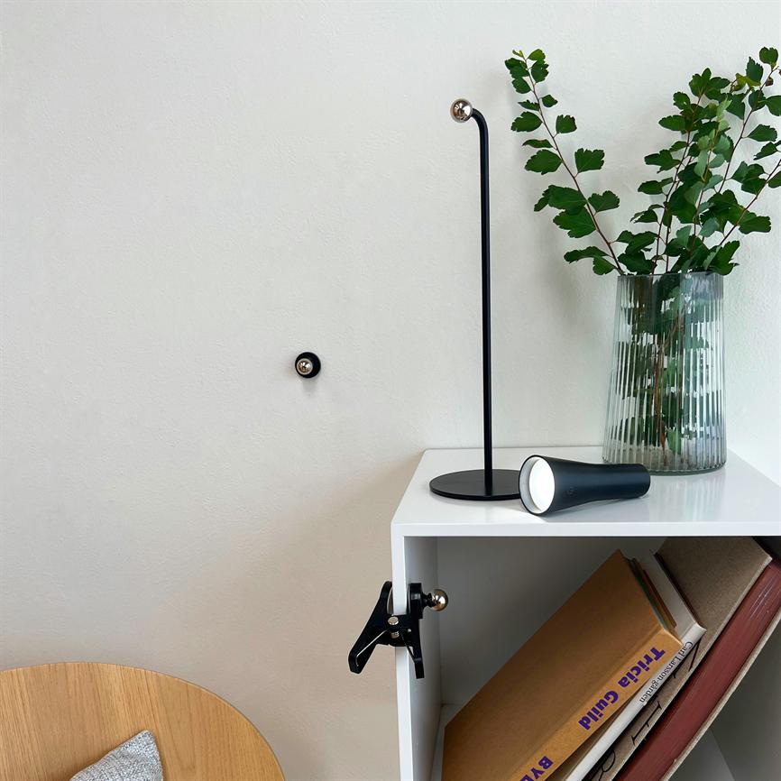 Sort bordlampe ligger på en hvid reol; en slank standerlampe står oprejst, ribbet glasvase med grønne grene ved siden af, bøger i reolen. Tekst: "Tricia Guild".