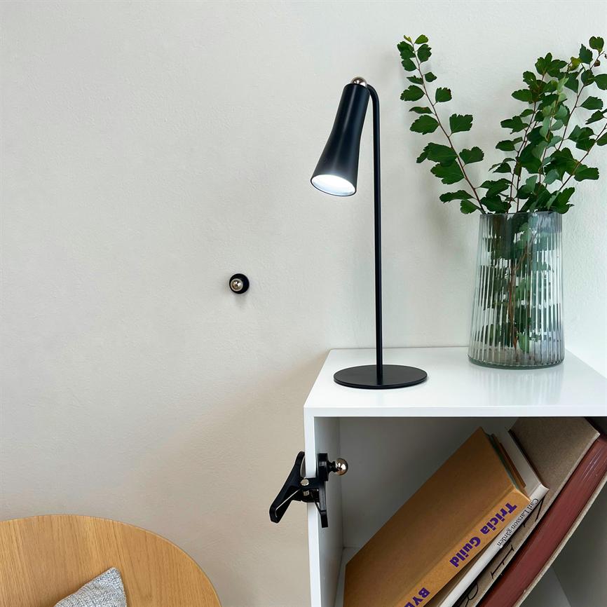En sort bordlampe lyser på en hvid hylde; hylden står ved en lys væg med en glasvase med grønne grene, og rummer bøger, herunder bogryg med teksten "Tricia Guild".