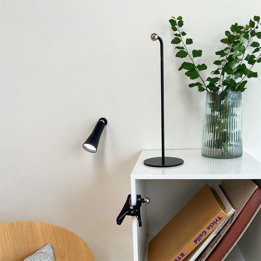 Sort væglampe lyser mod en lyst væg; en tynd sort bordlampe og en glasvase med grønne grene står på en hvid reol over skråstillede bøger. 
 Tekst på bogryg: "Tricia Guild"