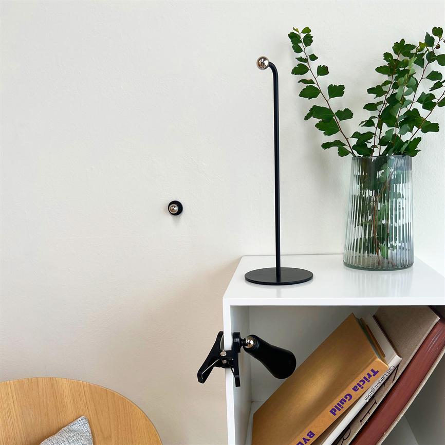 Sort, slank bordlampe står på en hvid hylde; den står ved en klar glasvase med grønne grene, over en åben reol med bøger. Tekst på bogrygge: "Tilda Guild".