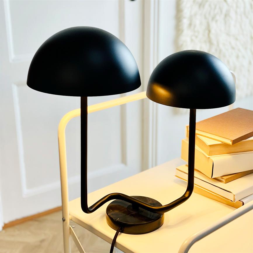 Sort dobbel bordlampe med to halvkugleformede skærme, der udsender varmt, gult lys; placeret på et hvidt rullebord ved en stak bøger, foran en dør i et lyst rum.