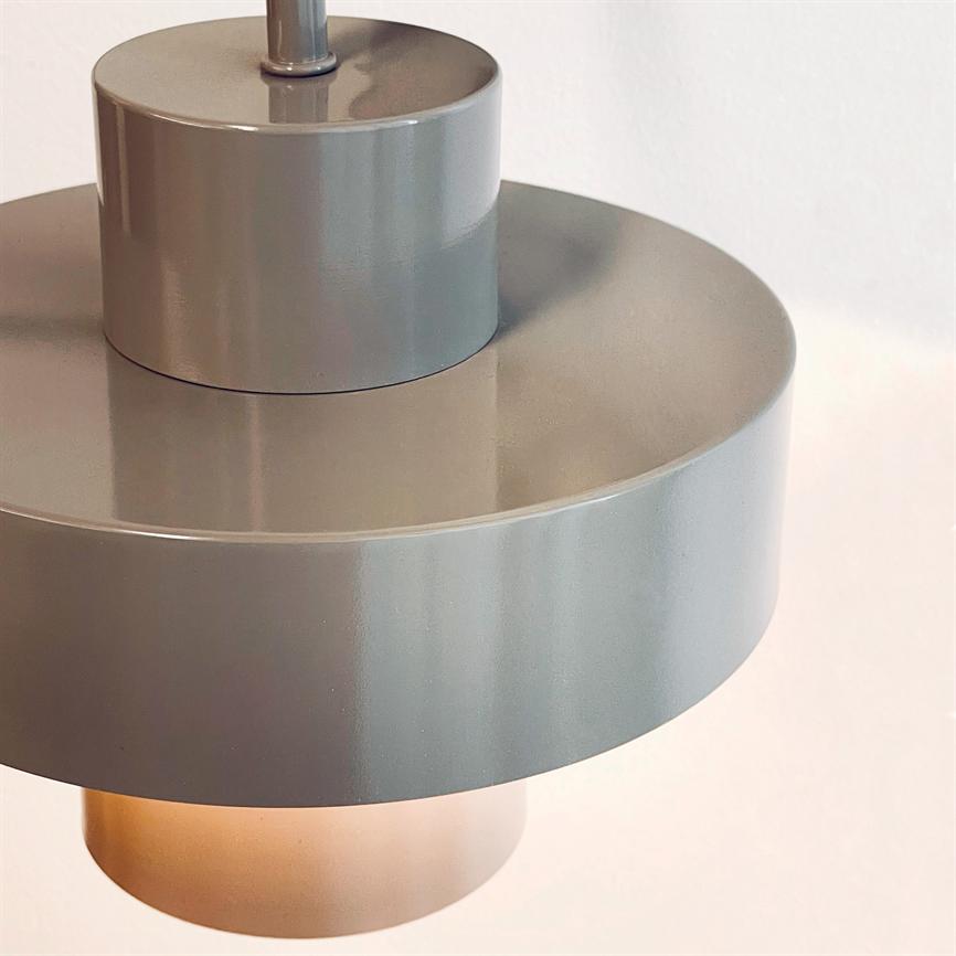En grå, rund pendellampe hænger og kaster blødt, varmt lys nedad; matt metaloverflade reflekterer svagt, med en neutral, lys væg som baggrund.