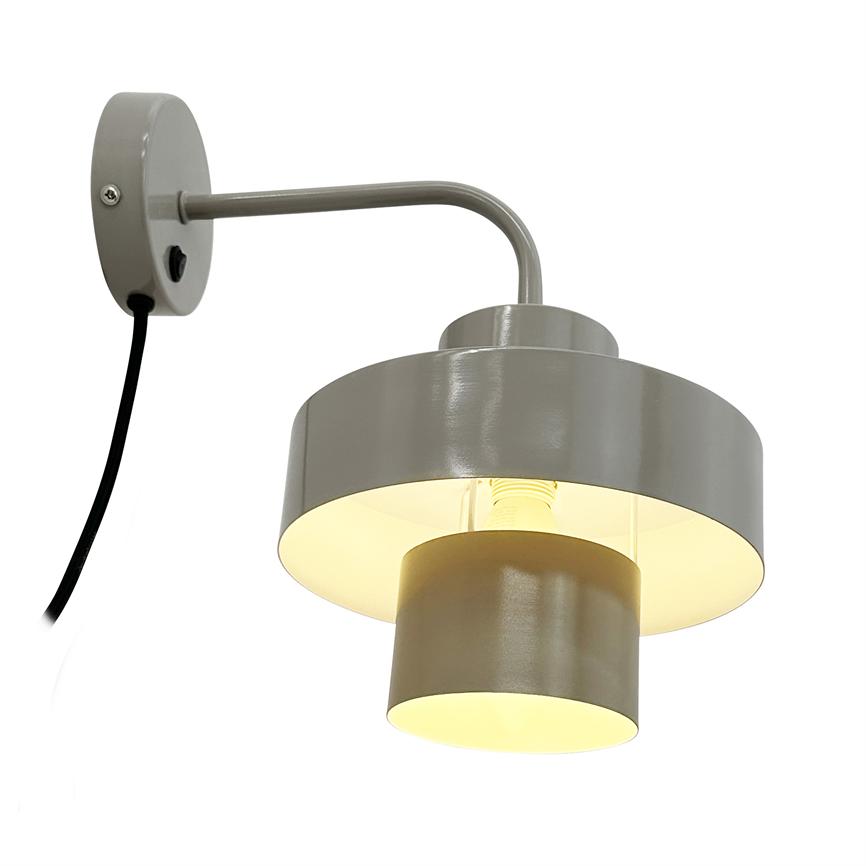 Væglampe monteret på en buet arm lyser varmt nedad; grå metallskærm i to niveauer, synligt sort kabel og rund vægbeslag mod neutral hvid baggrund.