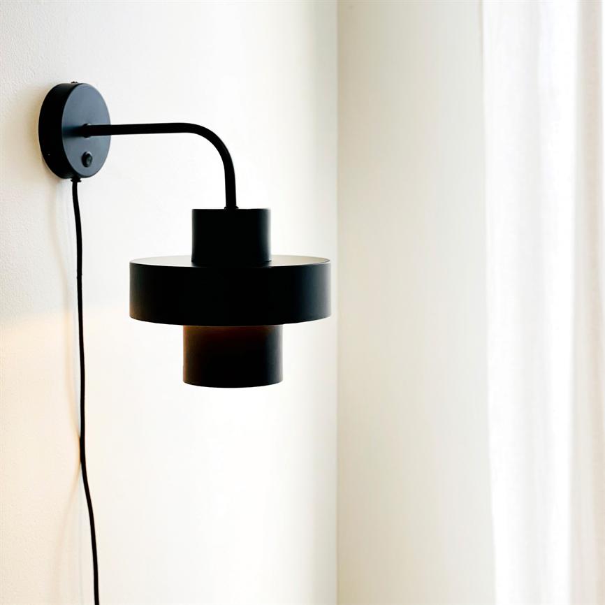 Sort væglampe hænger fra et buet armatur og kaster blødt lys på en lys væg ved siden af hvide gardiner i et minimalistisk, neutralt rum.