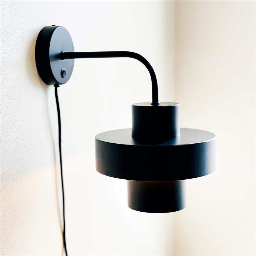 Sort, moderne væglampe med lagdelt cylindrisk skærm hænger fra en buet arm og rund vægmontering; ledning hænger ned langs en lys, neutral væg i et minimalistisk rum.