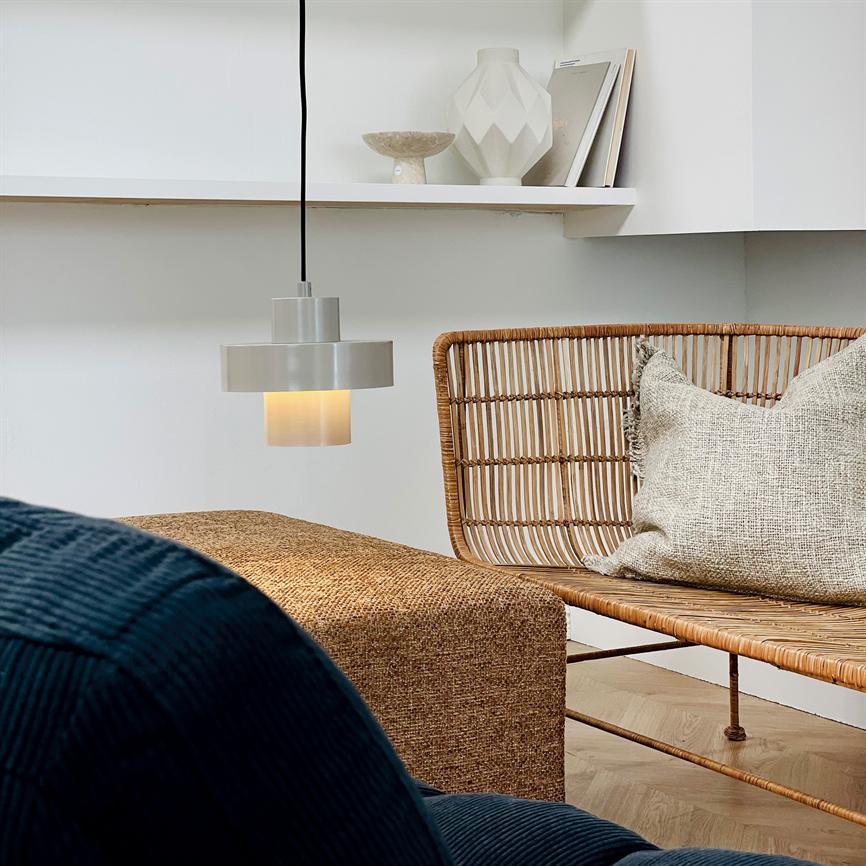 En matgrå hængelampe lyser varmt nedover i et lyst, minimalistisk hjørne med en rottingbænk, naturfarvet pude og en enkel hylde med keramiske vaser.
