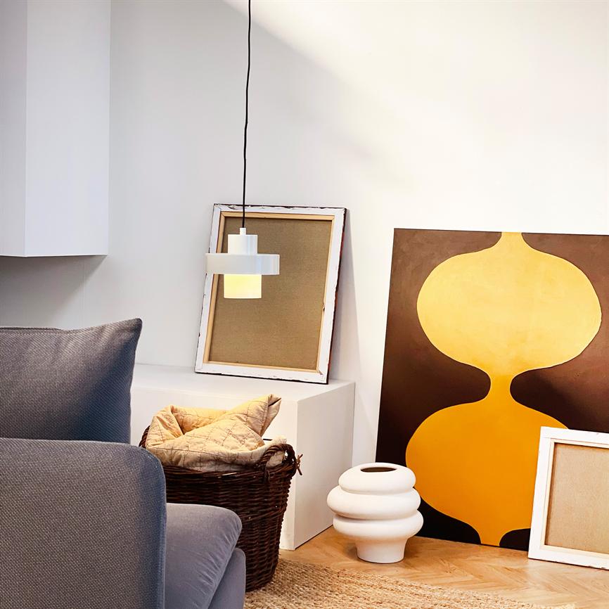 Hvid pendellampe hænger over en hvid skænk; foran står en kurv med puder, en hvid vase og et stort gulbrunt abstrakt maleri ved en grå sofa i et lyst, minimalistisk rum.