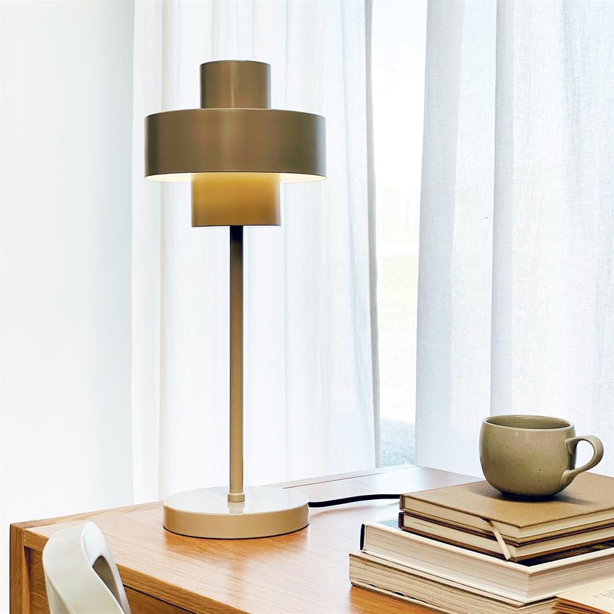 En moderne bordlampe lyser blødt og står på et træskrivbord ved et lyst vindue med hvide gardiner, ved siden af en stak bøger og en grøn keramikkop.