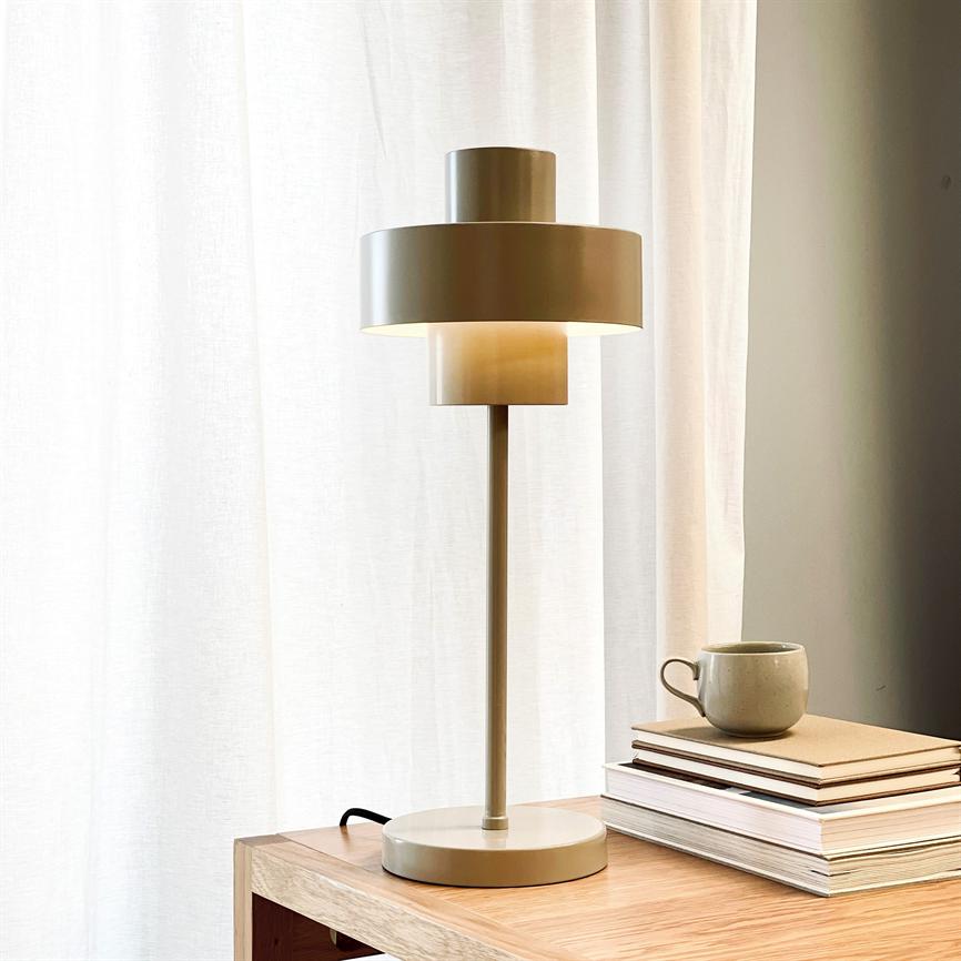 Bordlampe står tændt på et lyst træbord, udstråler blødt, varmt lys; ved siden af en stak notesbøger og en keramikkrus, foran et hvidt, solbelyst gardin i et roligt rum.