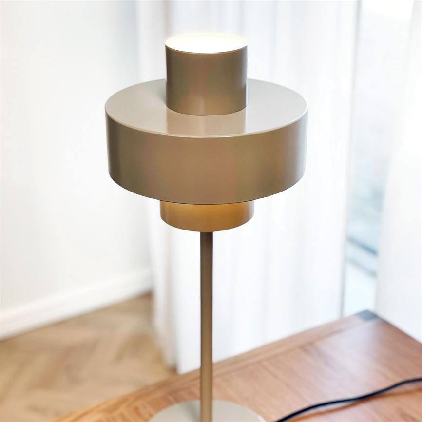 En moderne, metallisk bordlampe står og lyser blødt på et træbord foran lyse, gennemsigtige gardiner i et lyst rum med synligt trægulv.