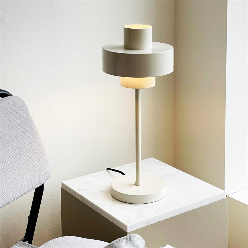 En beige bordlampe står og kaster varmt, diffust lys; på en marmoroverflade af en lav plint ved vindueskarmen, med en lys grå polstret stol til venstre.