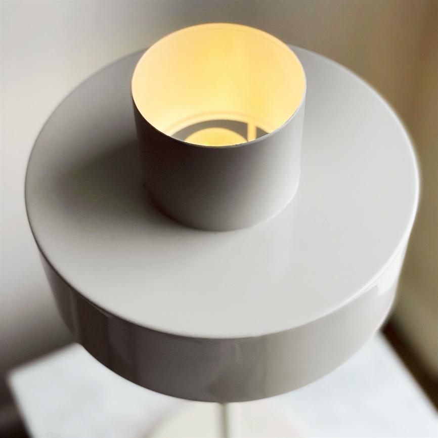 Hvid bordlampe afgiver varmt, dæmpet lys gennem en øverste cylindrisk åbning, placeret på en hvid bordplade i et lyst, neutralt indendørs rum.