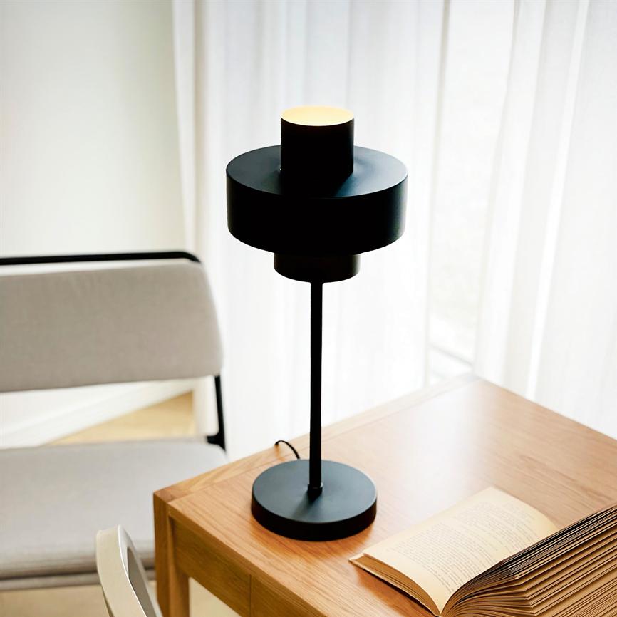 Sort bordlampe lyser svagt over en åben bog på et lyst træbord; en polstret stol står ved bordet, og hvide gardiner filtrerer dagslys i baggrunden. Bogtekst ikke læsbar.