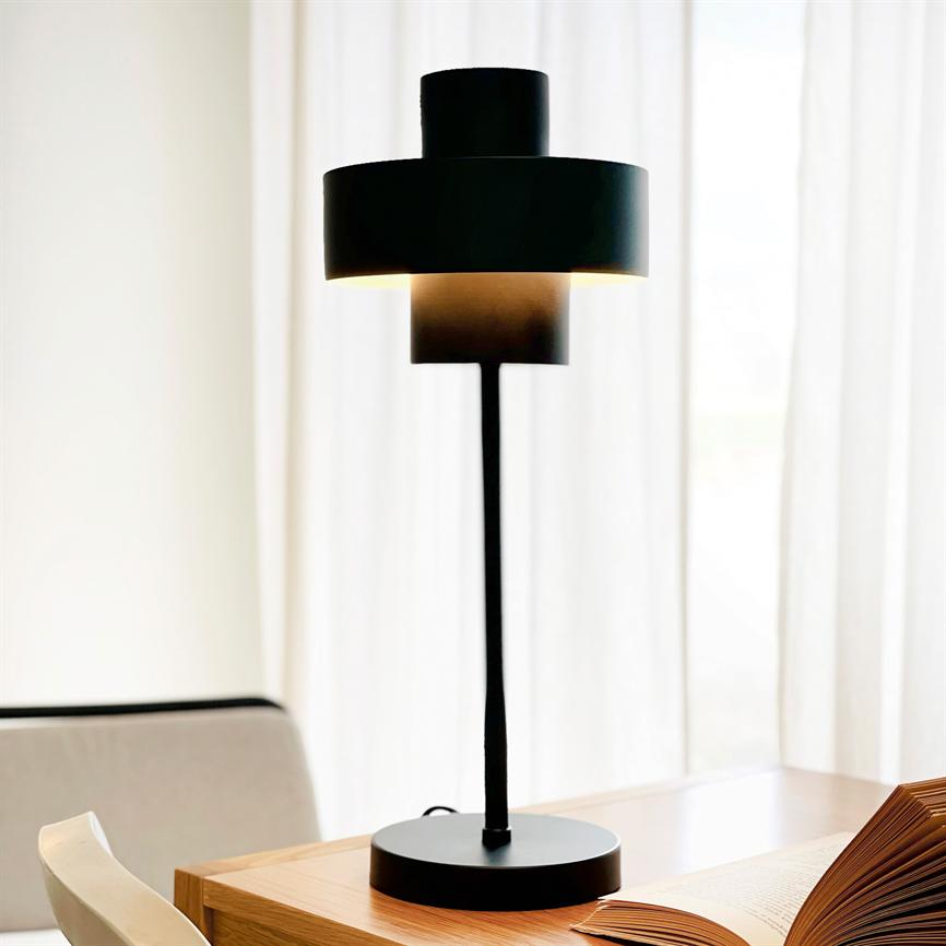 Sort moderne bordlampe står tændt og kaster varmt lys nedad; placeret på et træbord ved et åbent bogopslag foran lyse gardiner med en stol til venstre.