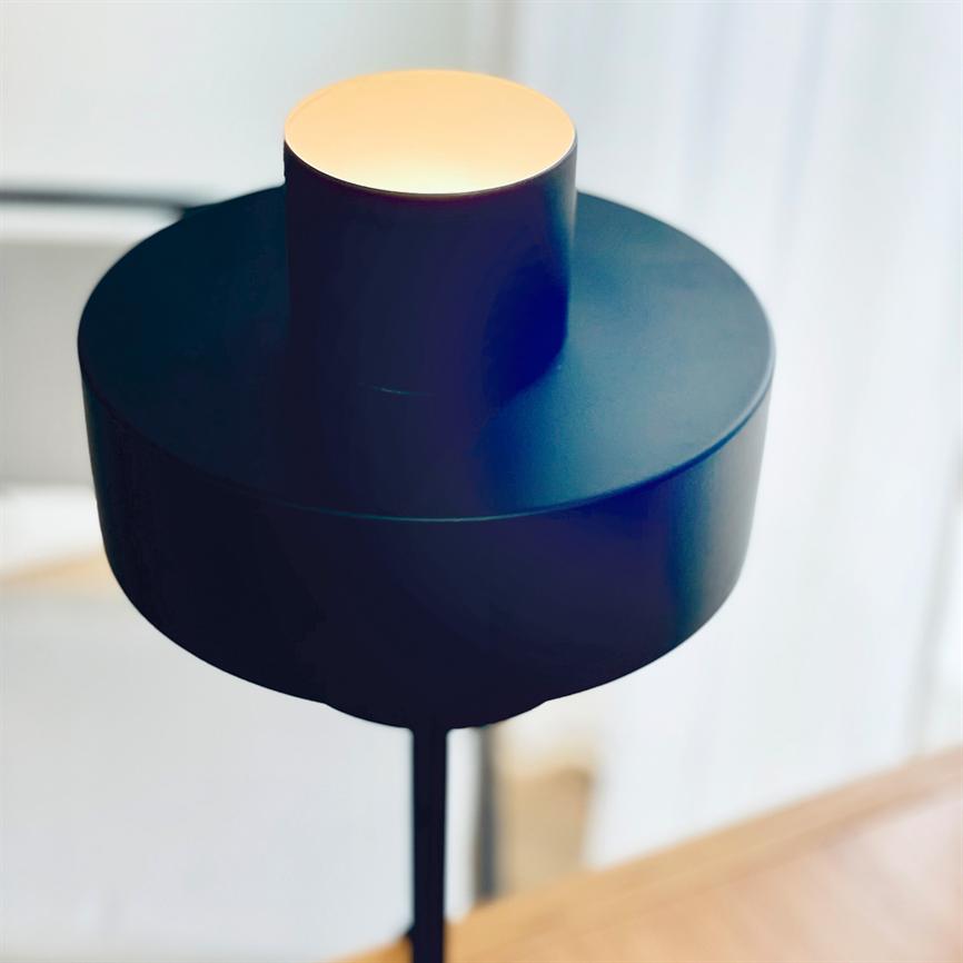 En moderne sort gulvlampe udstråler varmt lys fra en rund åbning i en cylindrisk skærm, stående ved et lyst vindue over et træbord i et minimalistisk rum.