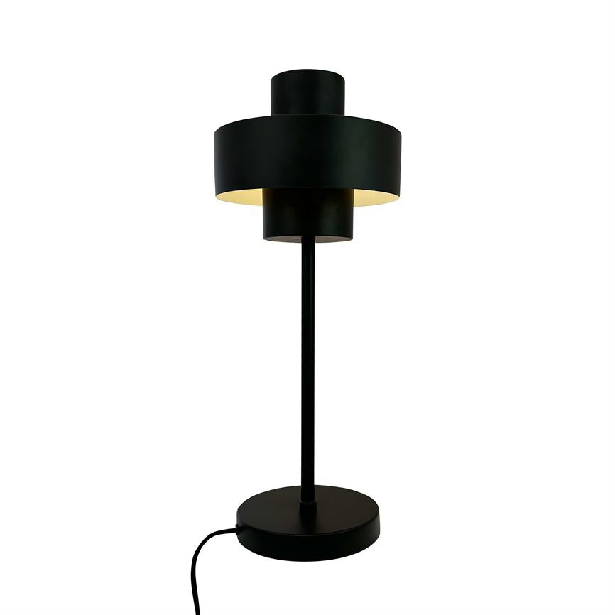 En sort bordlampe med overlappende cylindriske skærme udsender varmt, nedadrettet lys; den står på en rund fod med synlig ledning mod en ensartet hvid baggrund.
