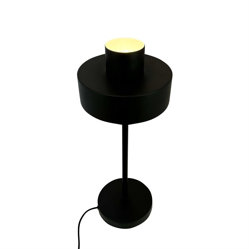 Sort bordlampe med cylindrisk skærm, udsender en varm opadrettet glød; tynd stang forbinder skærm til rund fod, synlig sort ledning, placeret mod en ensartet hvid baggrund.