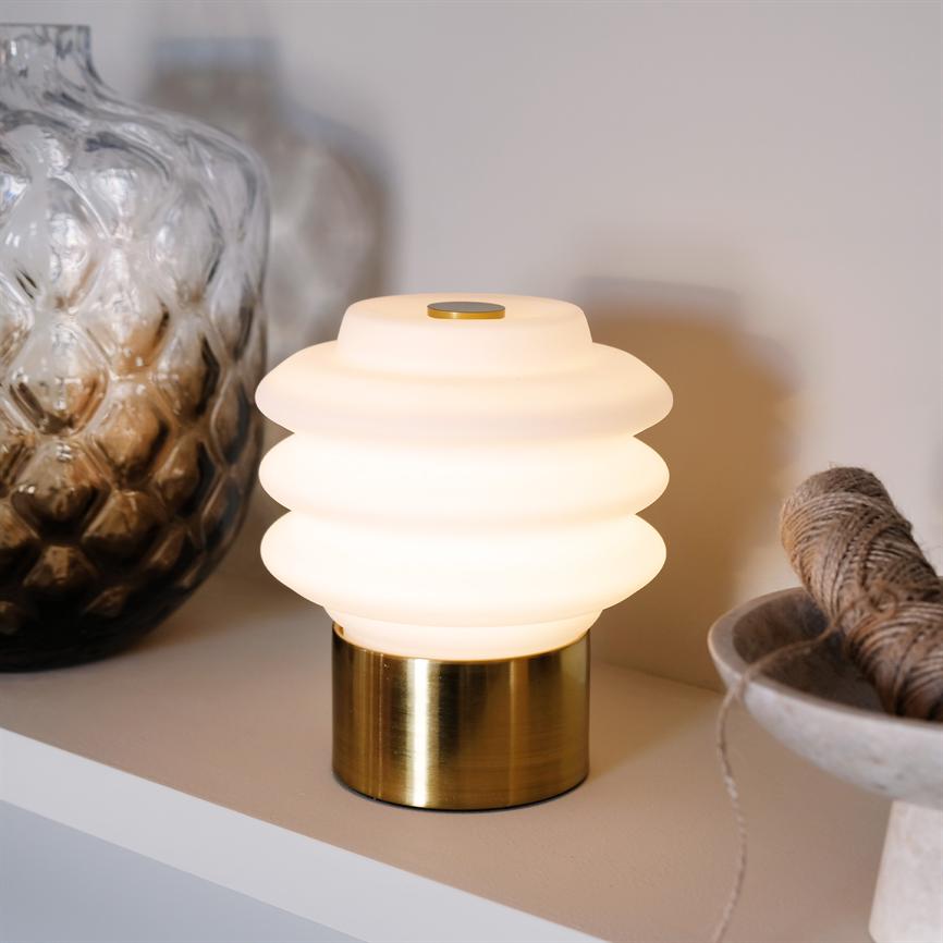 Bordlampe med ribbet opalglas og messingfod udsender blødt, varmt lys på en hylde ved en mønstret glasvase og en skål med snor.