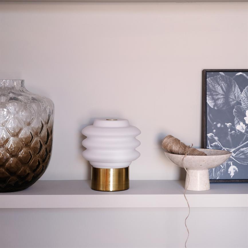 Hvid, bølget bordlampe på messingfod står på en beige hylde; til venstre ses en stor brunlig mønstret glaskrukke, til højre en skål med snor og et sort-hvidt plantefoto.