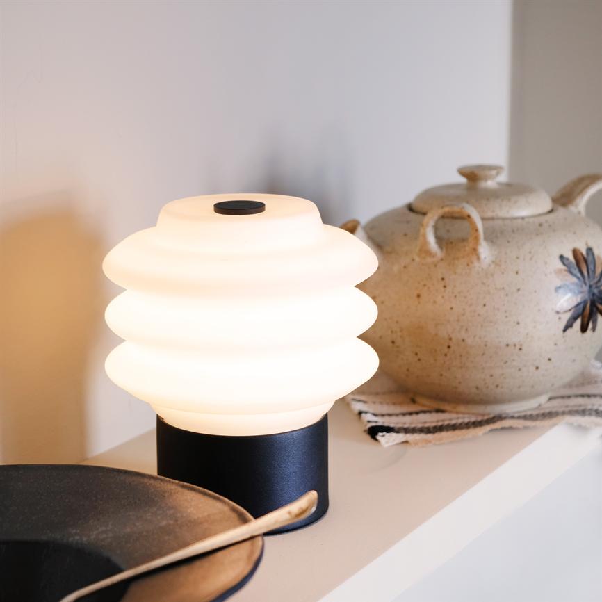 Bordlampe med ribbet, hvid glaskuppel afgiver varmt, blødt lys; står på en hylde ved en beige keramisk tekande og en mørk tallerken med en metalske i et roligt, indendørs køkkenmiljø.