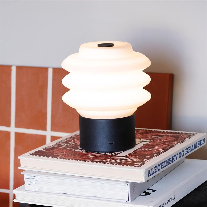 Lampe på bogstak: En hvid, bølget bordlampe lyser blødt og står på to kunstbøger ved et bord med terracottafliser i baggrunden. Tekst på bogryg: "ALECHINSKY OG BRAMSEN".