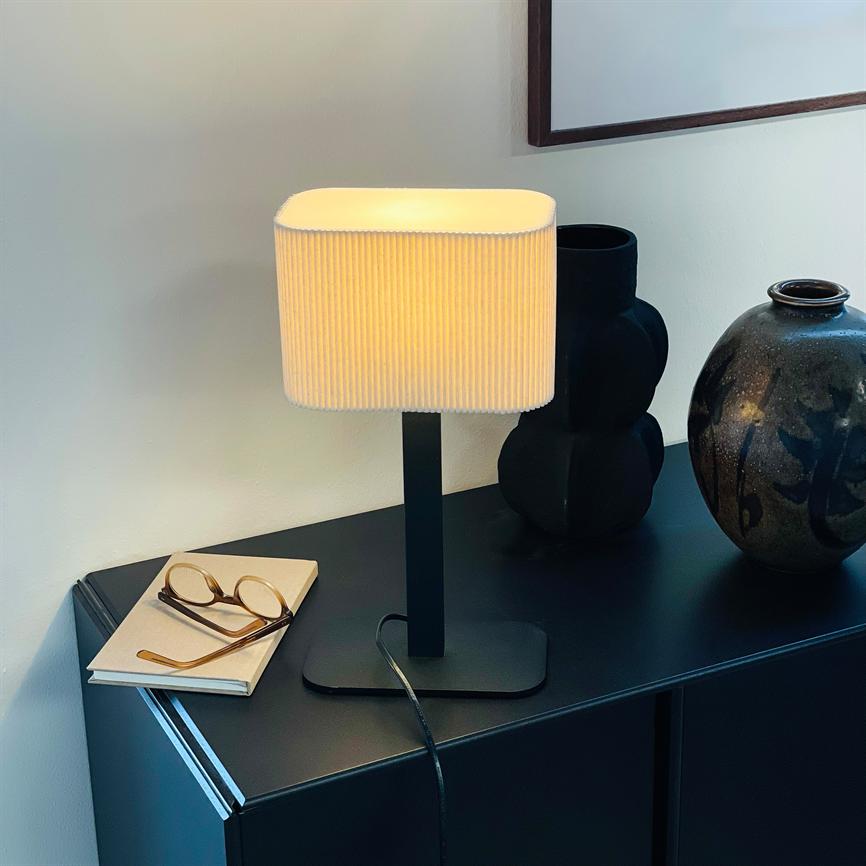 Bordlampe lyser blødt, stående på et mørkt skab ved siden af to dekorative vaser og et par briller på en notesbog, foran en hvid væg med indrammet billede.