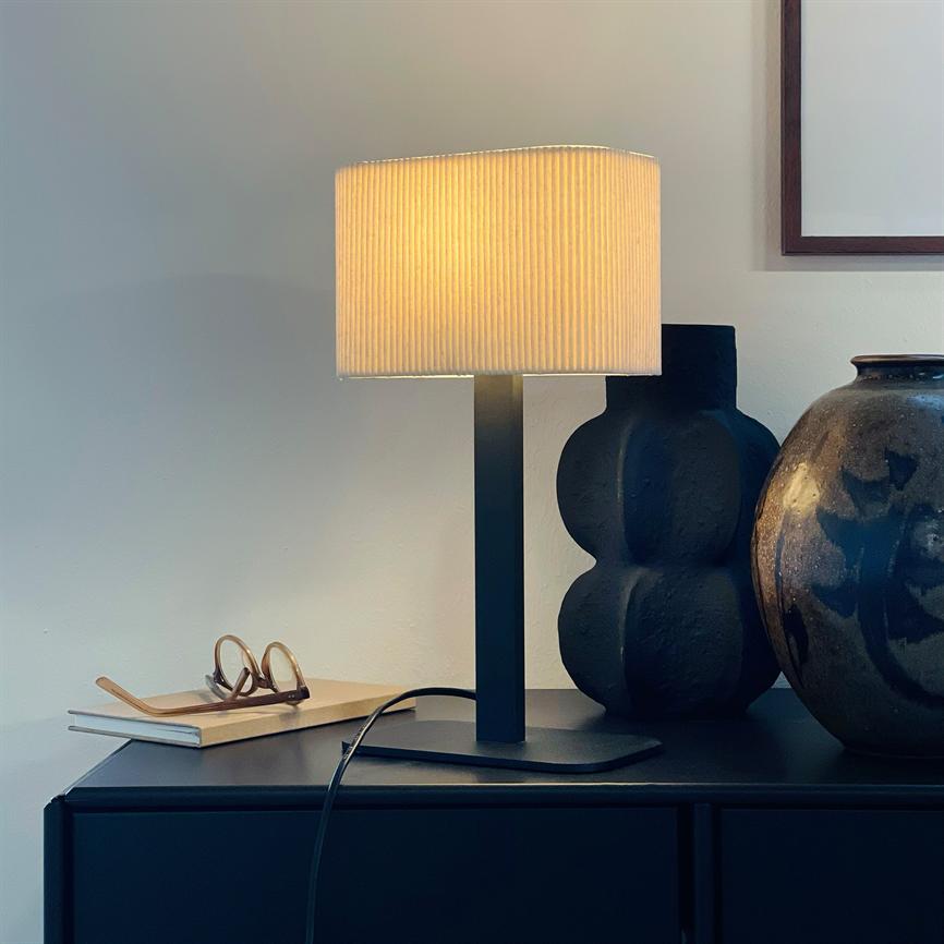 Bordlampe lyser varmt over et sort skab; ved siden står en bunke bøger med briller, en sort skulpturel vase og en brun, glaseret krukke mod en lys væg.
