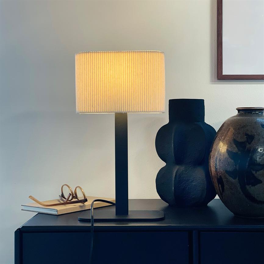 Bordlampe udsender varmt, gyldent lys; den står på et mørkt skab sammen med en sort vase, en glaseret krukke og et par briller på en notesbog under en indrammet plakat.