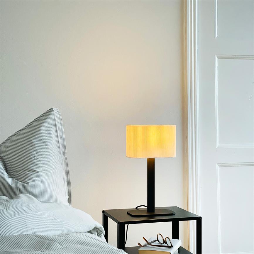 Bordlampe står tændt på et sort natbord, kaster varmt gult lys; ved siden af ligger briller og en bog, nær en stribet pude og en hvid dør i et lyst soveværelse.