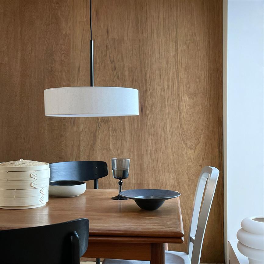 En hvid cylindrisk loftslampe hænger over et træspisebord; sorte stole, et gennemsigtigt vinglas, keramiske skåle og en bambusdamper står på bordet foran en varm trævæg.