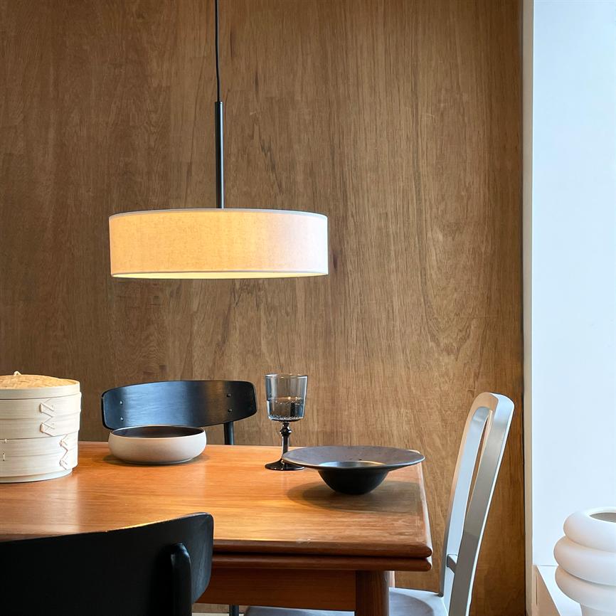En rund stoflampe hænger over et teakbord; to skåle og et vinglas står stille på bordet, omgivet af sorte og hvide stole foran en varm, træbeklædt væg.