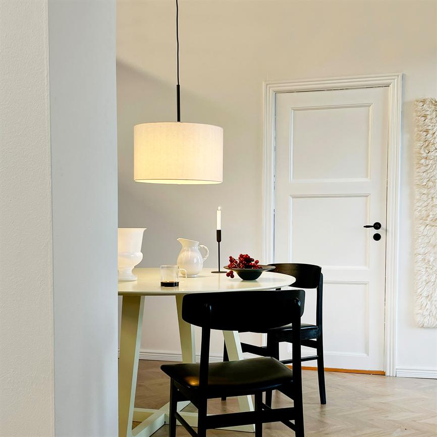 Et rundt spisebord står i rummet med to sorte stole; en hængelampe lyser over kande, lysestage og en skål med røde bær i en lys, minimalistisk spisestue ved en hvid dør.